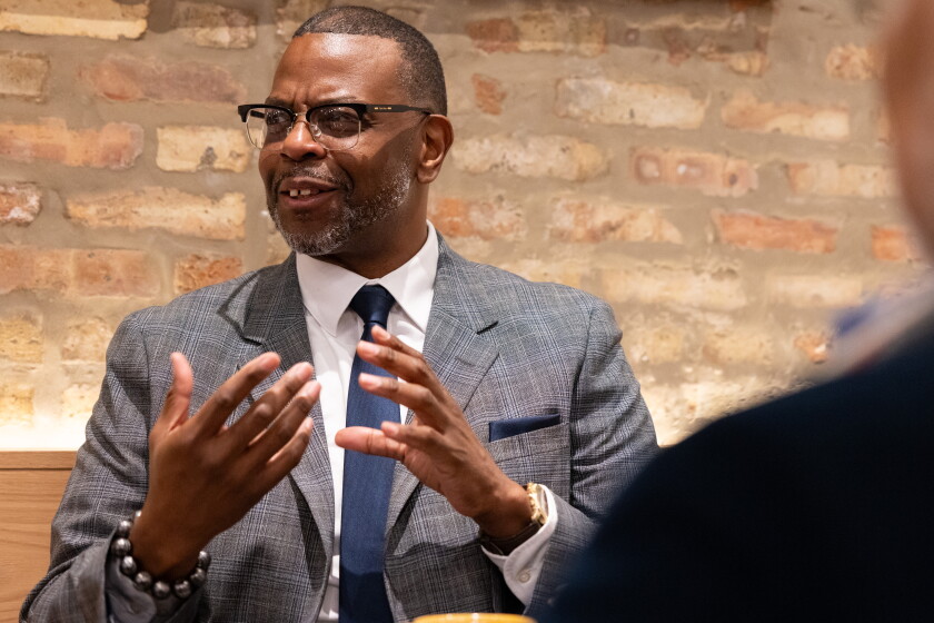 A Black man wearing glasses, a grey suit and a blue tie is seated in a cafe and speaking with others at his table.