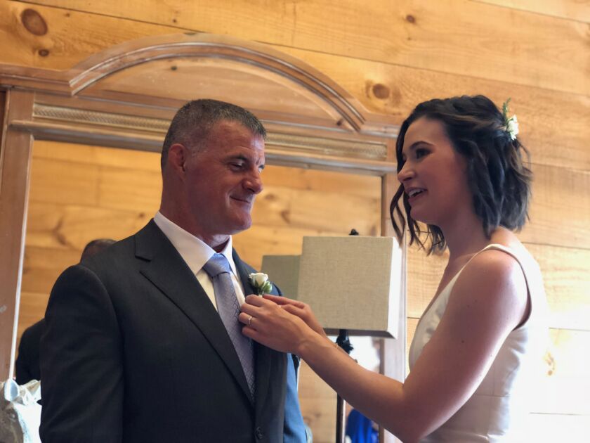 Rebecca Dougherty, wearing a white wedding gown, is pinning a rose onto the lapel of a jacket worn by her father, Dale Saran. Dougherty still hasn’t been able to resolve a debt she received after the VA claimed she didn’t qualify to receive her dad’s Post-9/11 GI Bill benefit.