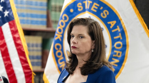 A woman is standing. There is an American flag to the left. Behind the woman is a flag that reads, "Cook County State’s Attorney."
