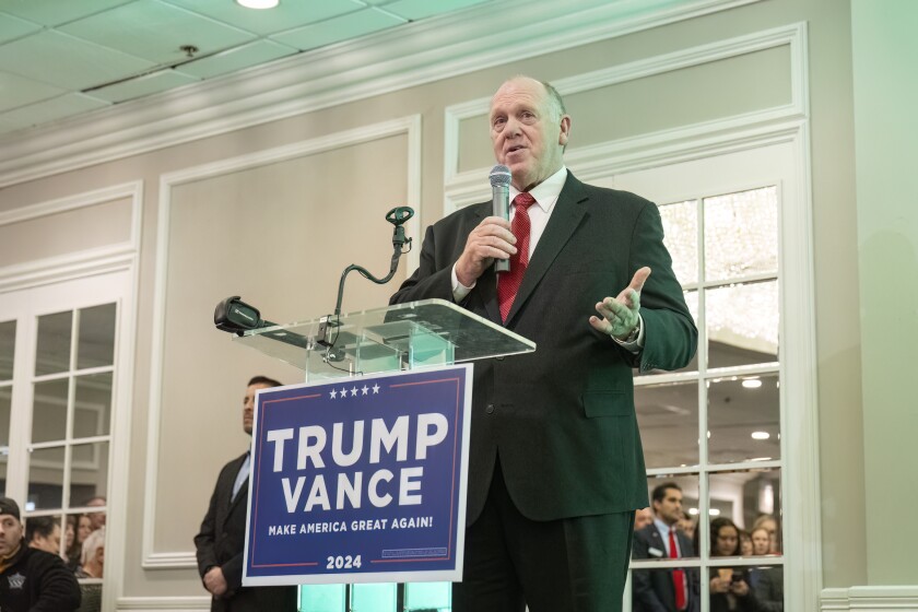 Tom Homan, wearing a black suit and red tie, holds a microphone while speaking at a clear acrylic podium hung with a blue "Trump Vance" sign, as the audience is reflected in the mirror behind him.